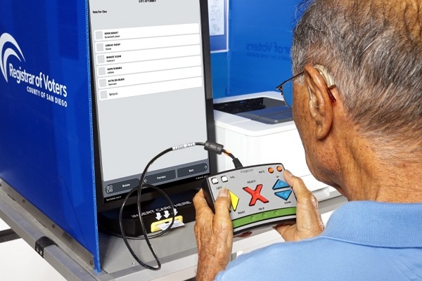 Man using hand-held controller (audio-tactile interface) to mark ballot choice on a ballot marking device screen